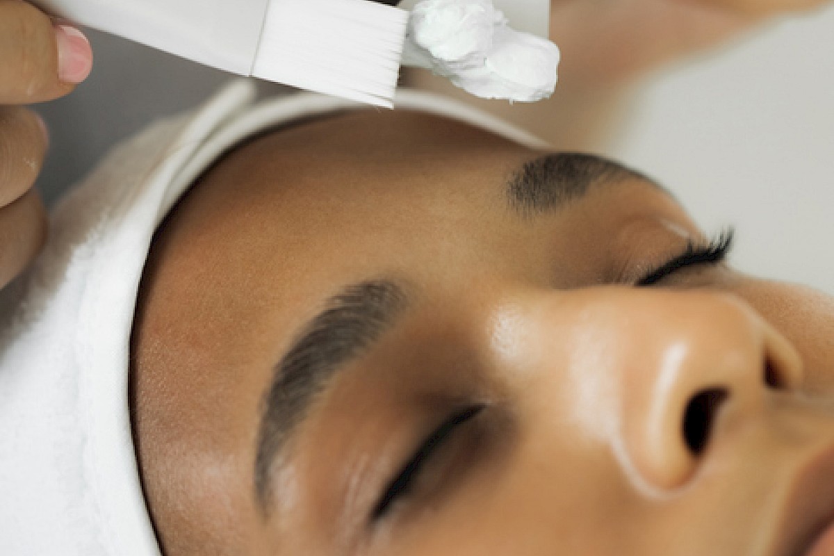 A woman is receiving a facial treatment with a white cream being applied using brushes.