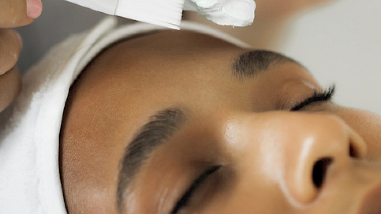 A woman is receiving a facial treatment with a white cream being applied using brushes.