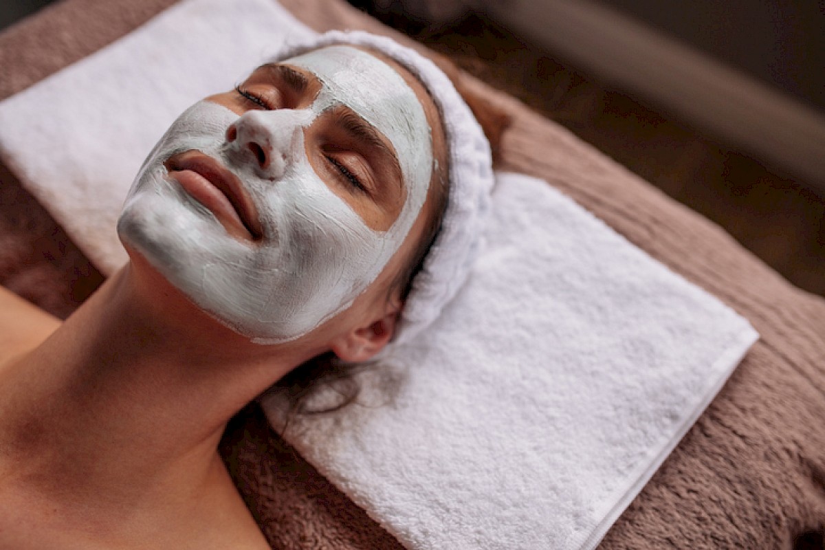 A woman is lying with eyes closed, wearing a facial mask in a spa or skincare treatment setting.