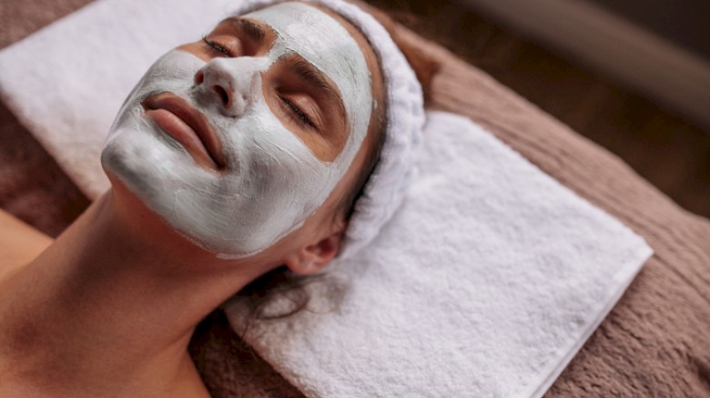 A woman is lying with eyes closed, wearing a facial mask in a spa or skincare treatment setting.