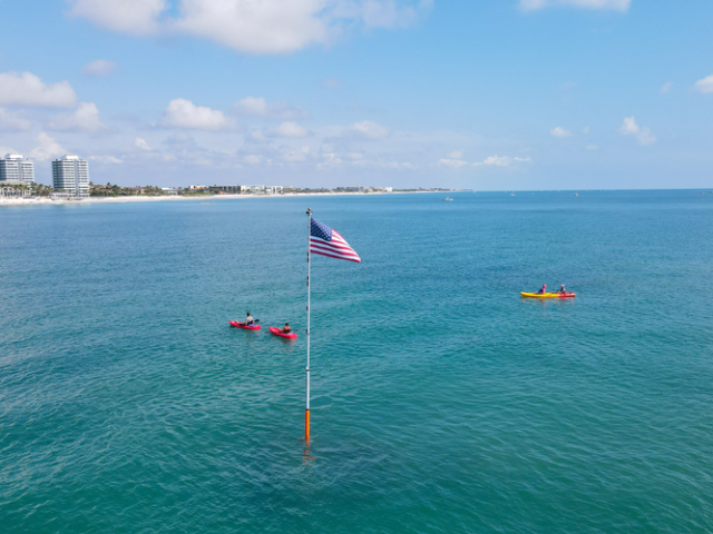Tres kayaks en el océano cerca de un alto mástil con la bandera estadounidense, edificios y una playa visibles a lo lejos bajo un cielo parcialmente nublado.