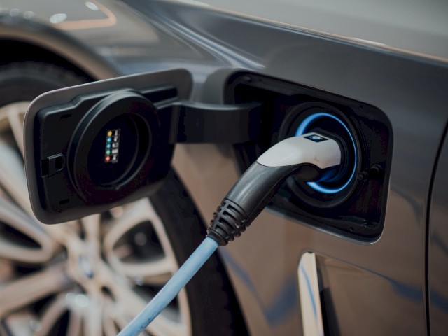 La imagen muestra un primer plano de un vehículo eléctrico cargándose en una estación de carga, con el enchufe conectado al puerto de carga del coche.