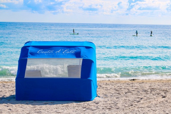 A blue beach tent on the sandy shore at Costa d'Este Beach Resort, with people paddleboarding on the ocean in the background.