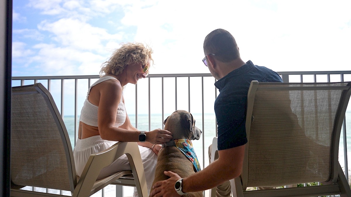 Two people are sitting on a balcony with a dog, enjoying the view of the ocean and sky.