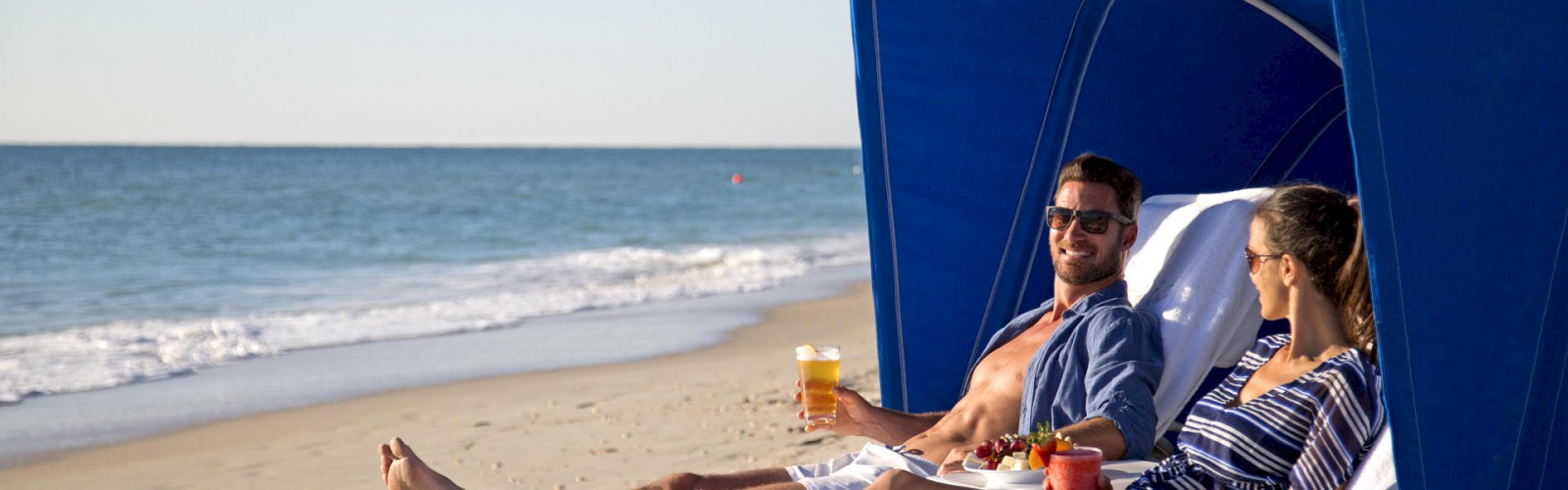 Two people relax on reclining chairs under a blue canopy at the beach, holding drinks and enjoying the sunny weather, facing the ocean.