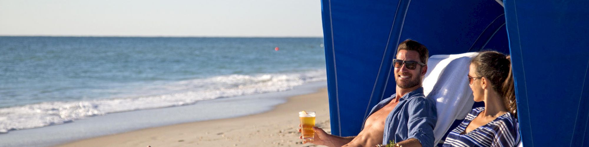 Two people relax on reclining chairs under a blue canopy at the beach, holding drinks and enjoying the sunny weather, facing the ocean.