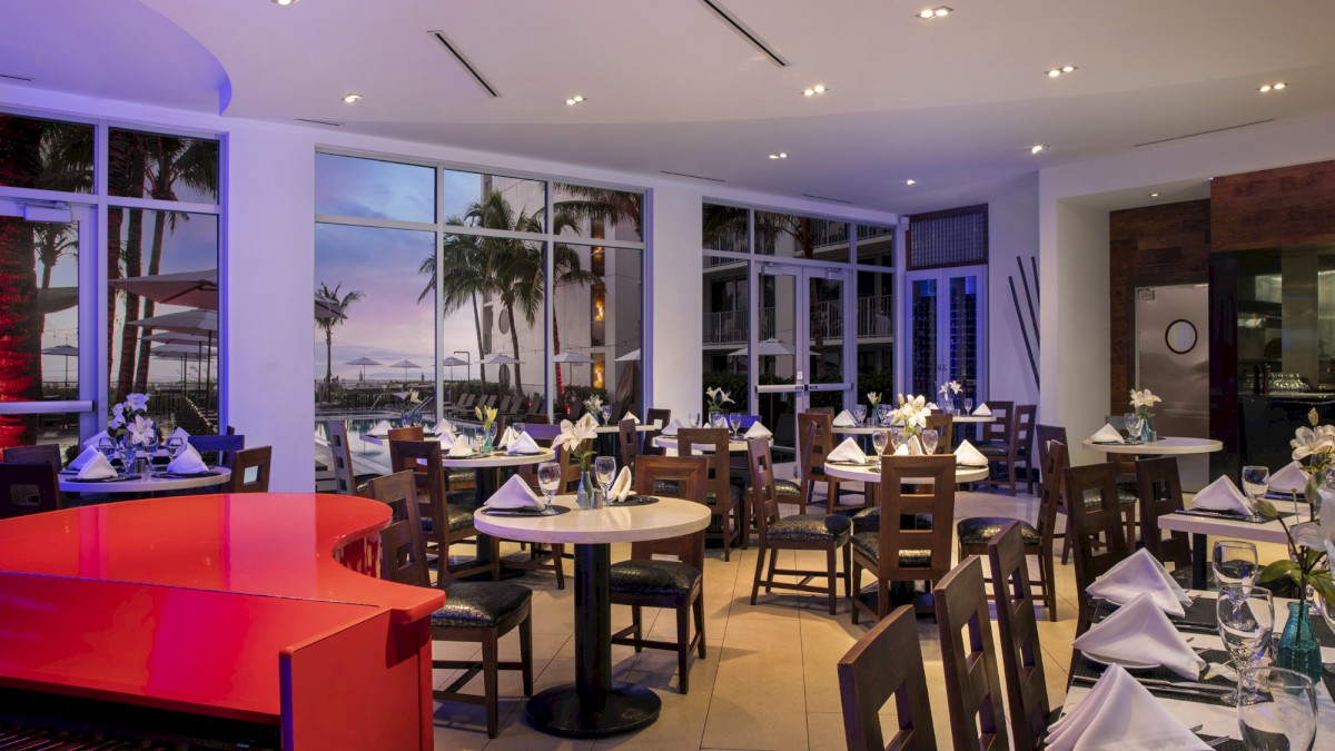 A cozy restaurant with elegantly set tables, a vivid red piano, and large windows revealing a palm-studded view of a sunset or twilight scene.