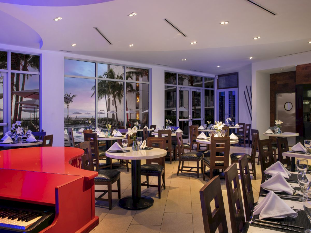A cozy restaurant with elegantly set tables, a vivid red piano, and large windows revealing a palm-studded view of a sunset or twilight scene.