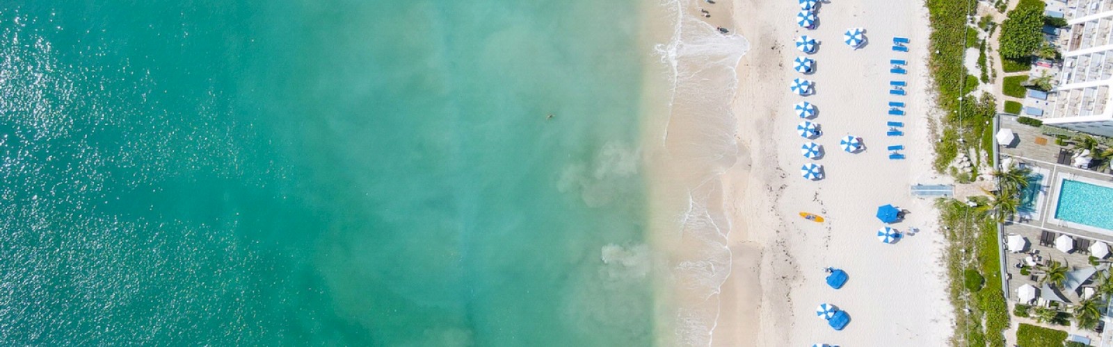 Aerial view of a beach with turquoise water, white sand, and blue umbrellas. Adjacent are buildings with pools and patches of green vegetation.