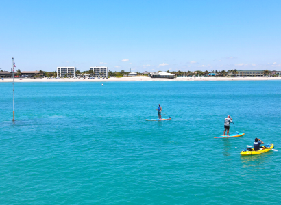 Three people are paddleboarding and kayaking in clear blue water near a shore with buildings in the background, under a clear sky.