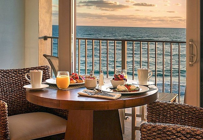 A cozy dining area with a table set for breakfast overlooks a serene ocean view through a sliding glass door during sunset.