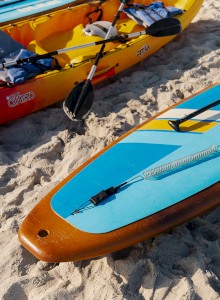 The image shows two kayaks and a paddleboard with paddles, life vests, and gear placed on a sandy beach, ready for use.