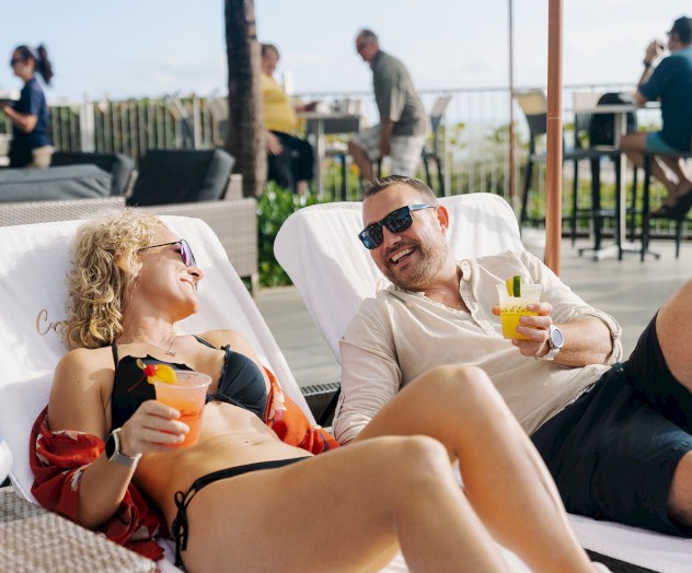 A man and woman relaxing on lounge chairs, smiling and holding drinks, with other people in the background near a bar or patio area.