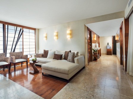 A modern and spacious living area with a large sofa, chairs, wooden table, wall lights, and adjoining corridor.