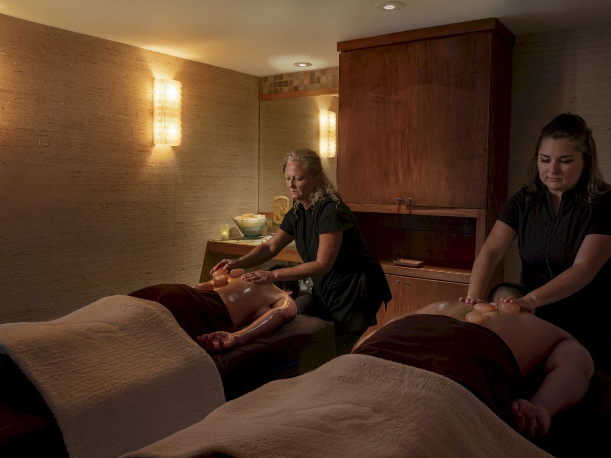 Two individuals are receiving massages from two therapists in a dimly lit room with warm lighting and cozy ambiance.