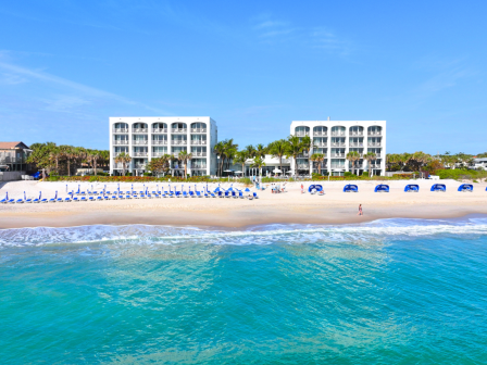 The image shows a beachfront view with two modern hotel buildings, lined beach chairs and umbrellas, and calm turquoise water under a clear blue sky.