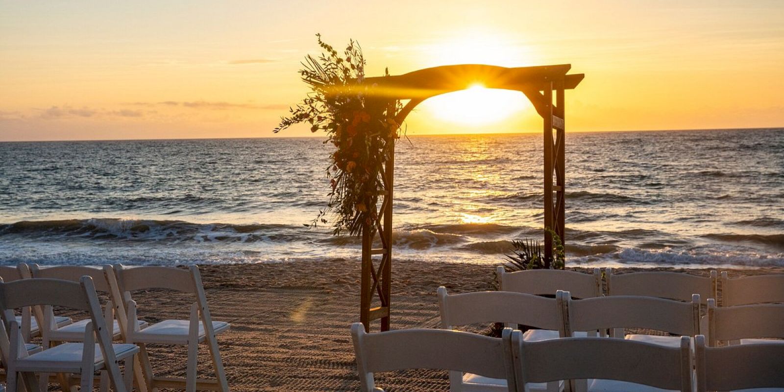 The image shows a beautiful beachside setup for a wedding or event, with a decorated archway and rows of chairs facing the ocean at sunset.