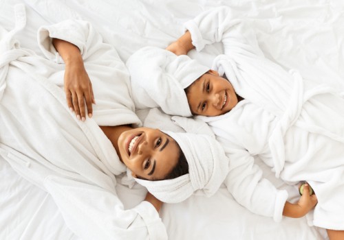 A happy woman and young girl relax in towels and robes, enjoying a cozy, fun moment together on a white bed.