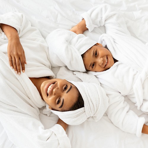 A happy woman and young girl relax in towels and robes, enjoying a cozy, fun moment together on a white bed.