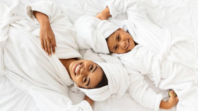A happy woman and young girl relax in towels and robes, enjoying a cozy, fun moment together on a white bed.