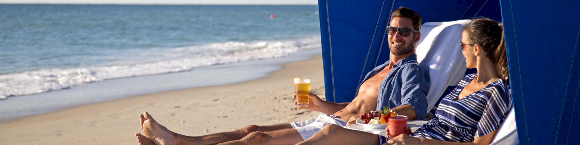 A man and a woman relax on lounge chairs under a blue beach canopy, enjoying drinks by the seaside on a sunny day.