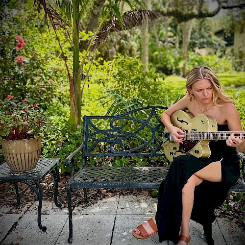 A young woman is sitting on a black garden bench playing an electric guitar in a lush, green outdoor setting with flowers and trees.