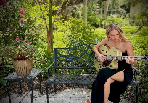 A young woman is sitting on a black garden bench playing an electric guitar in a lush, green outdoor setting with flowers and trees.