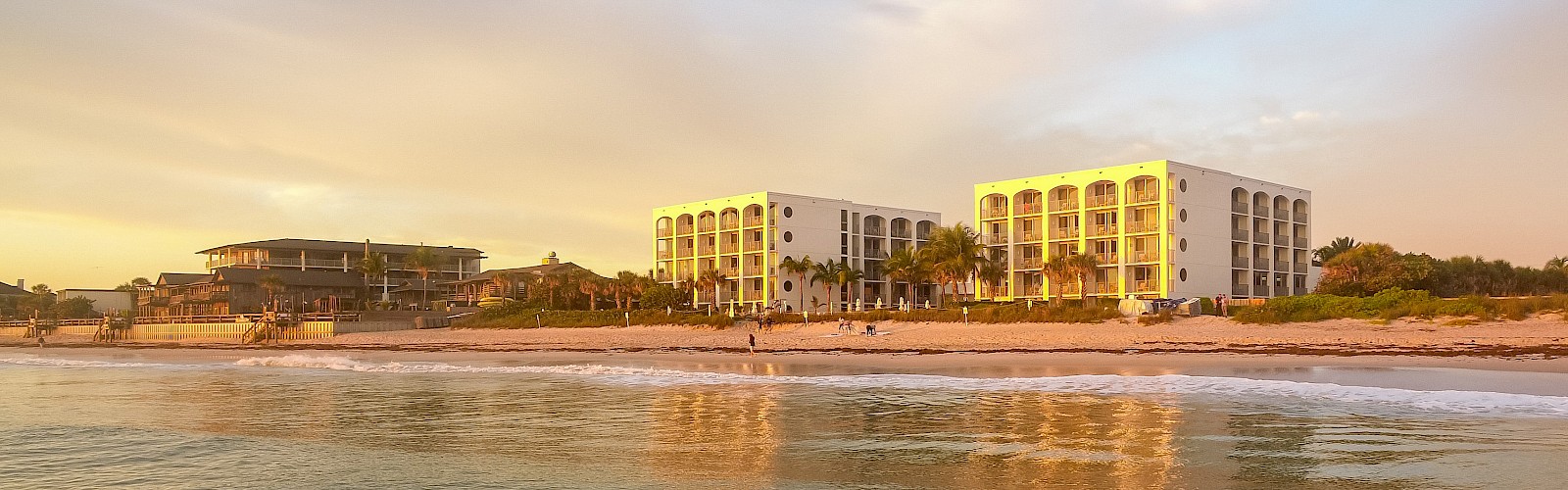 This image shows a serene beach scene at sunset with modern buildings and palm trees along the shoreline.
