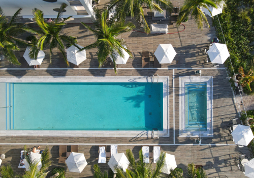 An aerial view of a swimming pool area with white umbrellas, palm trees, lounge chairs, and lush greenery surrounding a wooden deck.