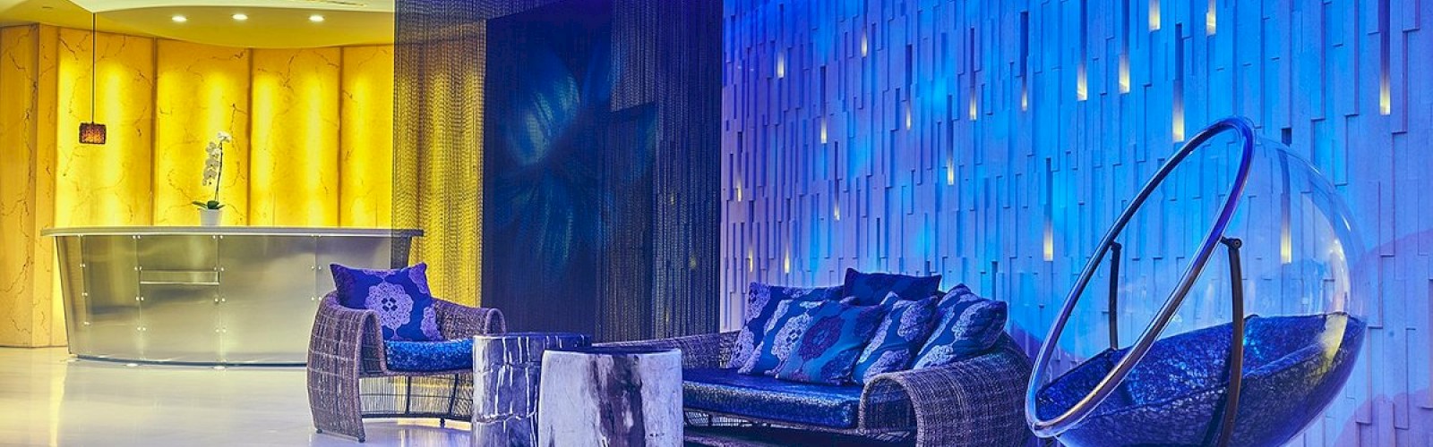 The image shows a modern lounge area with blue ambient lighting, wicker furniture, a glass bubble chair, and a reception desk in the background.
