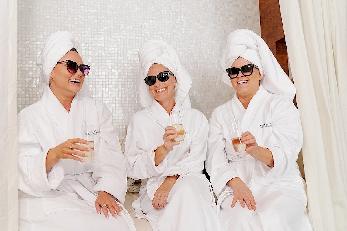 Three women in white robes and towels on their heads, wearing sunglasses, smiling, and holding glasses of champagne or sparkling beverage.