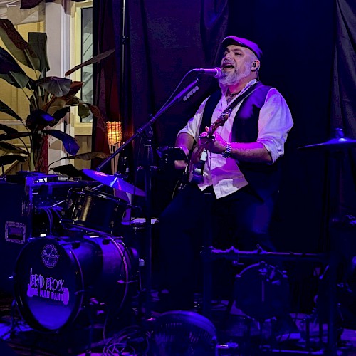 A musician with a beard, wearing a cap and vest, performs passionately singing and playing guitar on stage with drums and plants nearby.