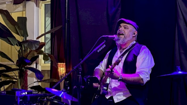 A musician with a beard, wearing a cap and vest, performs passionately singing and playing guitar on stage with drums and plants nearby.