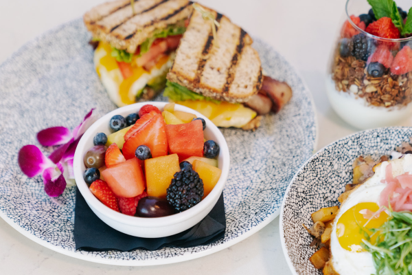 The image shows a meal with a sandwich, a bowl of mixed fruit, a yogurt parfait with berries, and a plate with fried eggs.