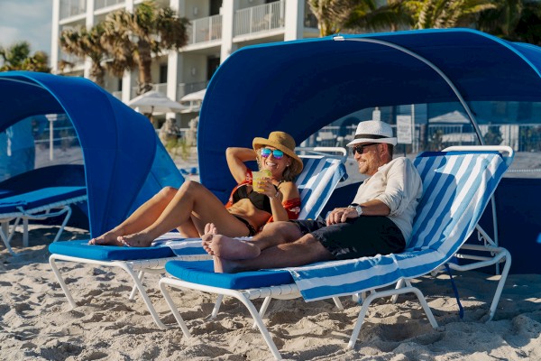 Two people relaxing on lounge chairs under blue cabanas at a beach, wearing hats and sunglasses, with a building and palm trees behind them.