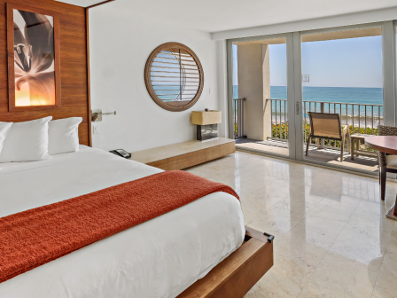 A hotel room with a large bed, ocean view balcony, and modern decor featuring wood accents and a tropical vibe.