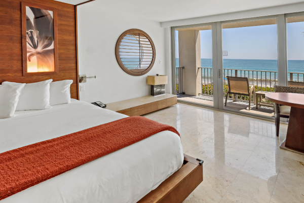 A hotel room with a large bed, ocean view balcony, and modern decor featuring wood accents and a tropical vibe.