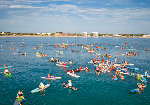 People paddleboarding and kayaking in a large group on a body of water, with buildings visible in the background under a clear sky.