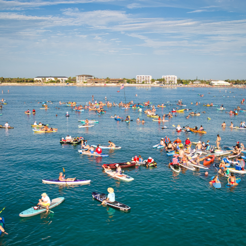 People paddleboarding and kayaking in a large group on a body of water, with buildings visible in the background under a clear sky.