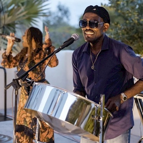 A musician smiling while playing steel drum, with a female singer performing in the background at an outdoor event.