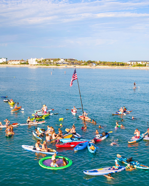 A group of people are on paddleboards and inflatables in the ocean, with an American flag on a pole, near a distant shoreline.