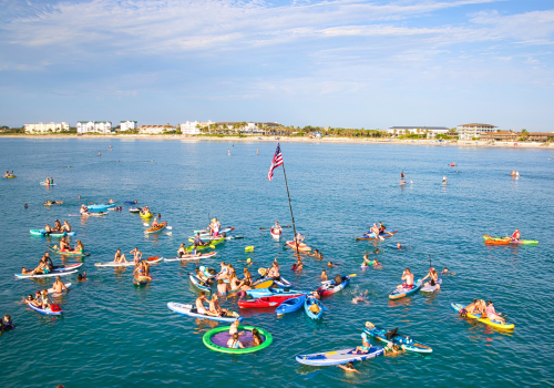 A group of people are on paddleboards and inflatables in the ocean, with an American flag on a pole, near a distant shoreline.
