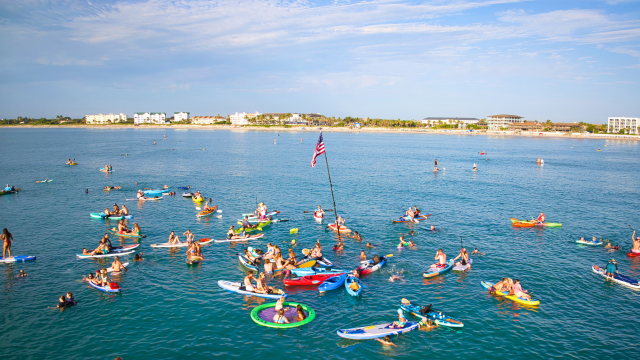 A group of people are on paddleboards and inflatables in the ocean, with an American flag on a pole, near a distant shoreline.
