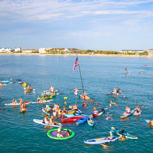 A group of people are on paddleboards and inflatables in the ocean, with an American flag on a pole, near a distant shoreline.