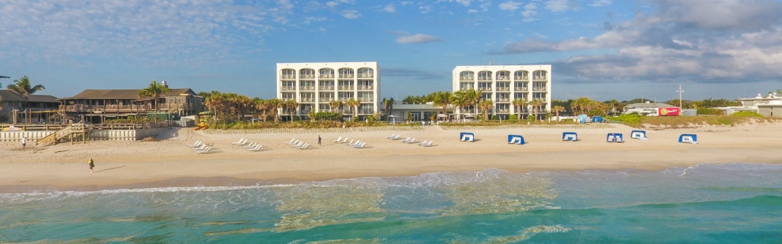 A beach scene with two white buildings, lounge chairs, umbrellas, and a few people near the shoreline under a partly cloudy sky.