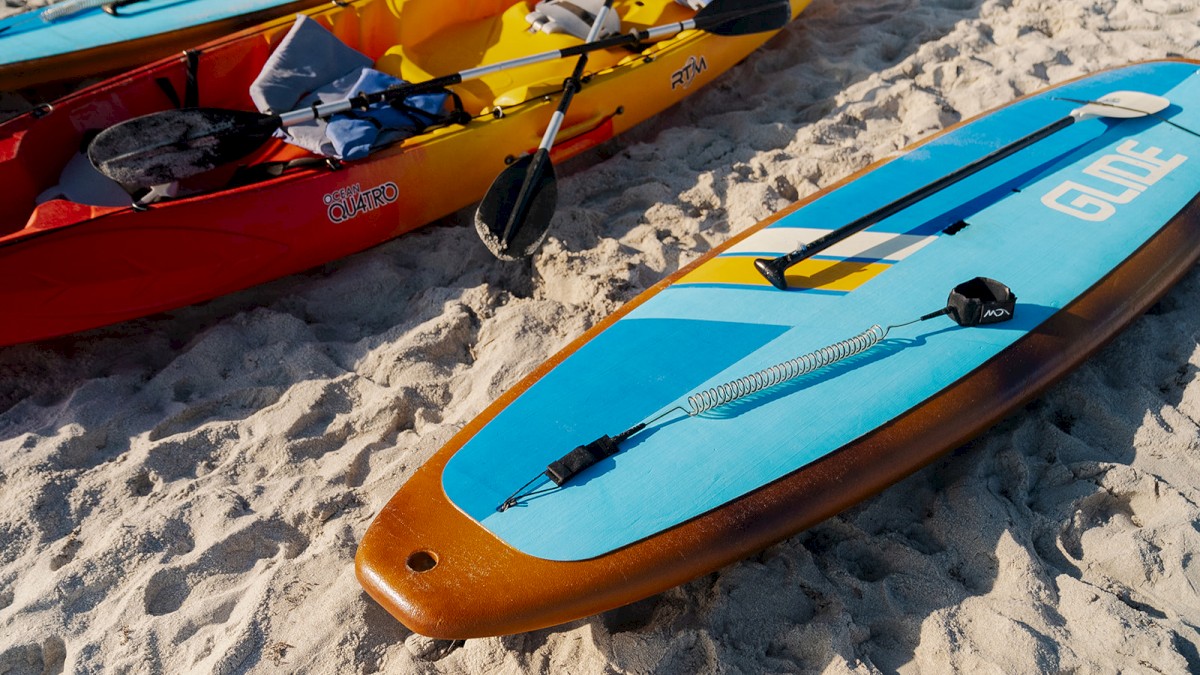 The image shows two colorful kayaks and a blue paddleboard on sandy ground, equipped with paddles and life vests for water activities.