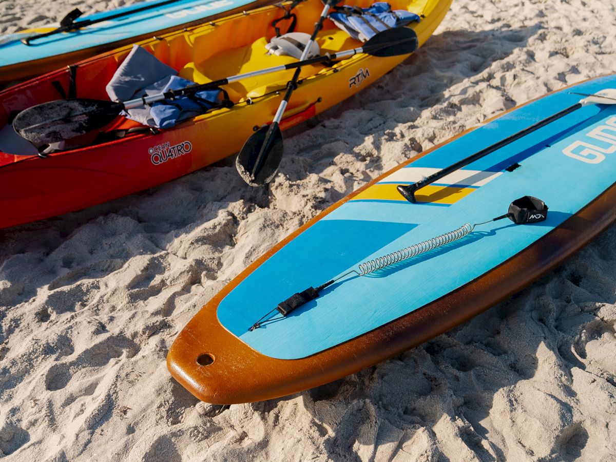 The image shows two colorful kayaks and a blue paddleboard on sandy ground, equipped with paddles and life vests for water activities.