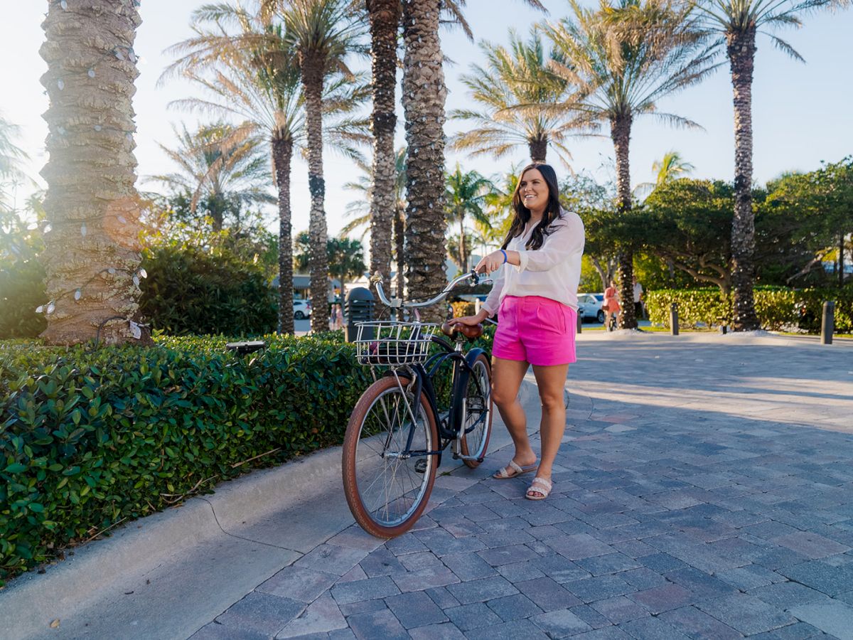 A woman stands beside her bicycle on a sunny day, surrounded by palm trees, wearing pink shorts and a white top, smiling and looking content.