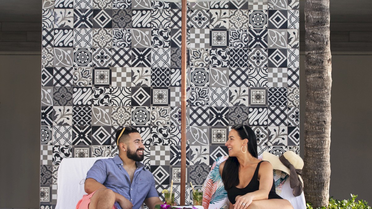 Two people are relaxing on lounge chairs by a pool, under an umbrella, with a patterned wall in the background. The scene is sunny and relaxed.
