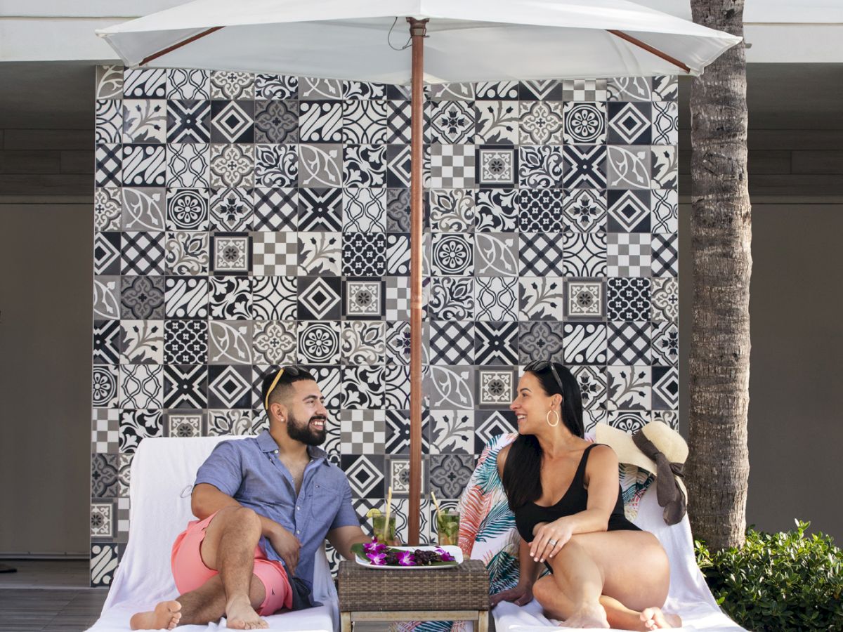 Two people are relaxing on lounge chairs by a pool, under an umbrella, with a patterned wall in the background. The scene is sunny and relaxed.